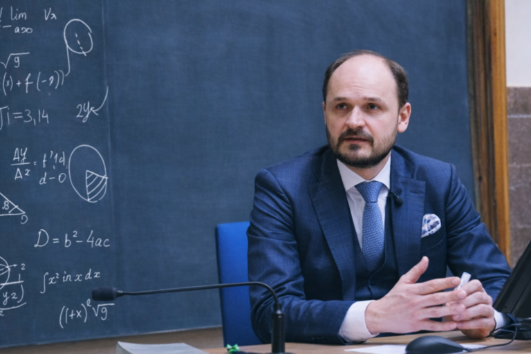 Muž v obleku sedí u stolu před tabulí s vzorci a diskutuje během odborné přednášky