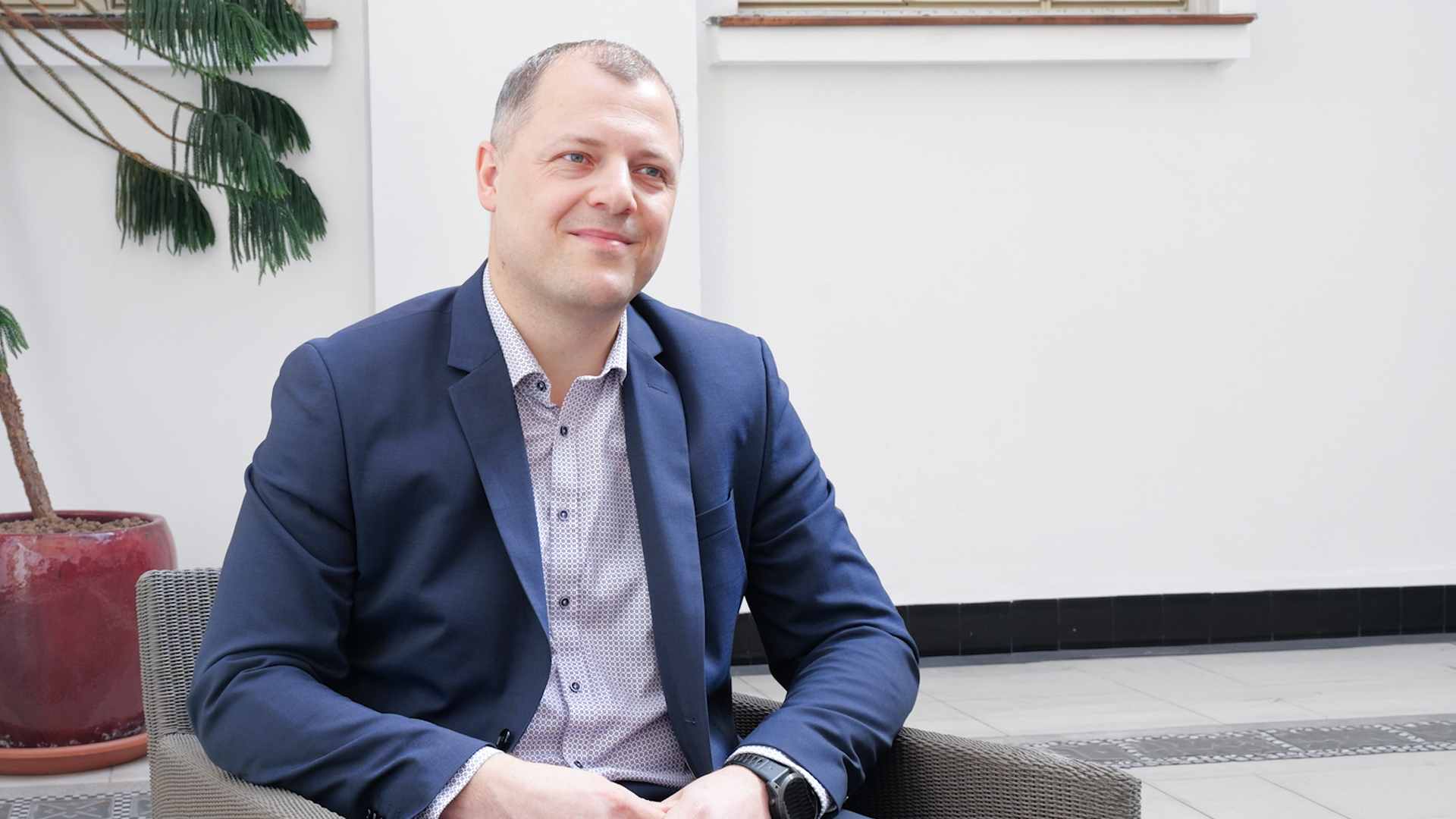 Muž v modrém saku sedí v křesle v moderním interiéru a během rozhovoru se usmívá. V pozadí je světlá hala s pokojovou rostlinou.