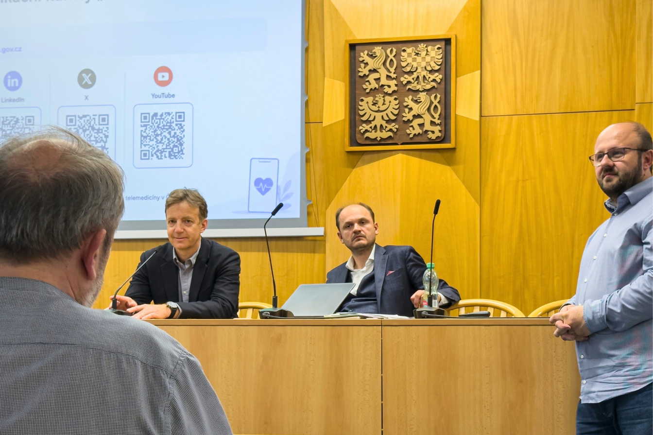 Odborný seminář k telemedicíně v jednacím sále Ministerstva zdravotnictví ČR. U řečnického stolu sedí přednášející, v popředí je vidět účastník akce a na projekčním plátně jsou zobrazeny QR kódy k online kanálům.